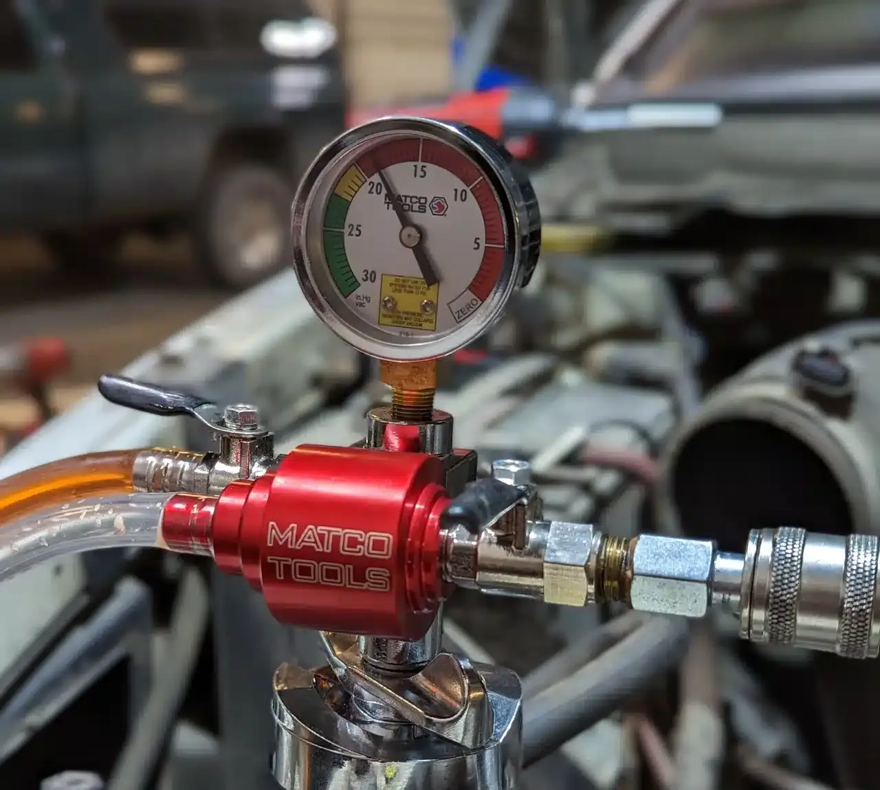 1023 Diesel & Fleet: A pressure gauge from Matco Tools is attached to a red valve, indicating the vacuum level in a car engine. The gauge shows a needle within a colored scale, and the vehicle’s engine components are visible in the background.