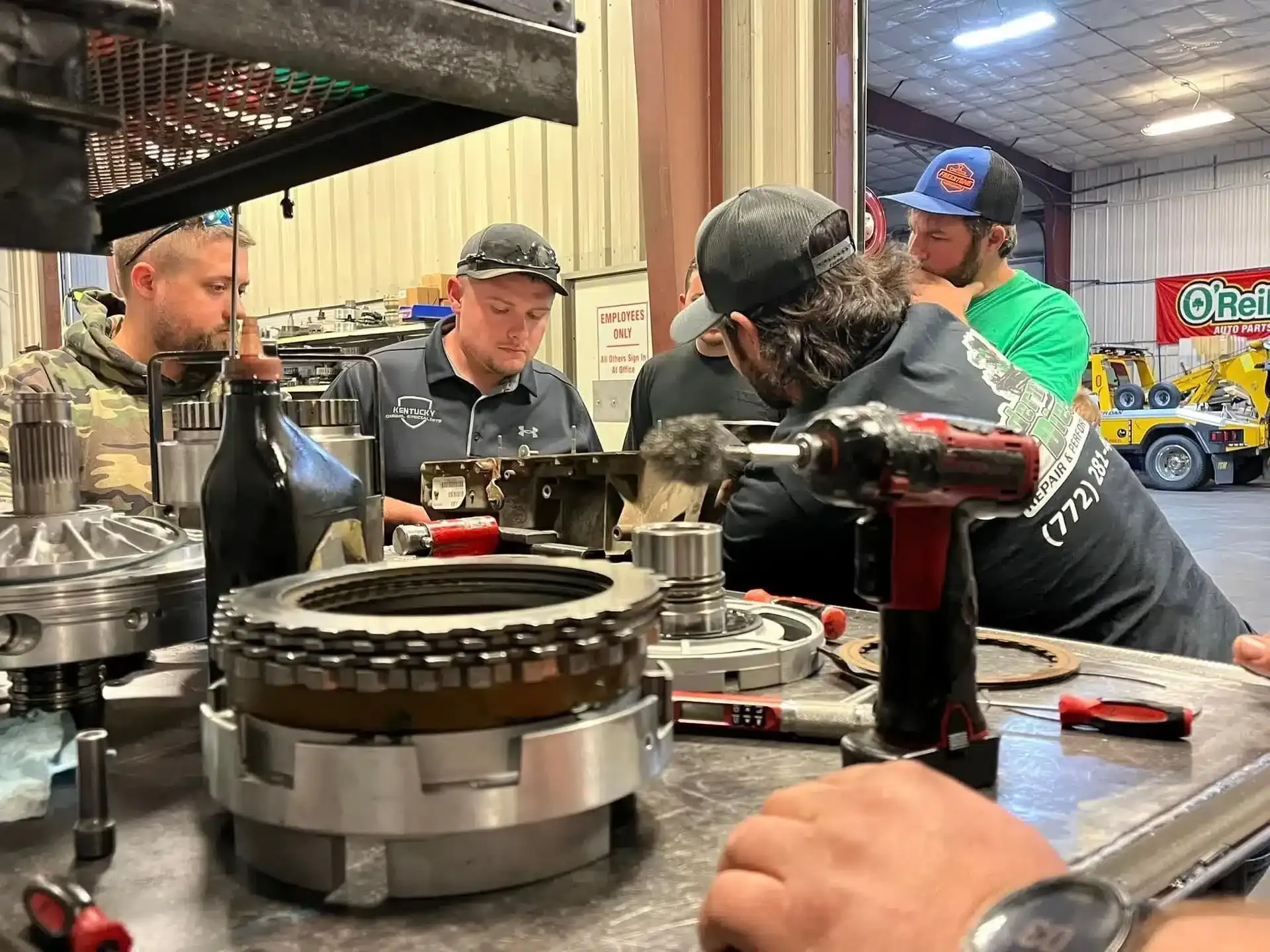 A group of people working on automotive parts at a workshop table, with tools and equipment scattered around. A man in a black cap is focused on a component, while others observe and discuss. Workshop interior and equipment are visible in the background.