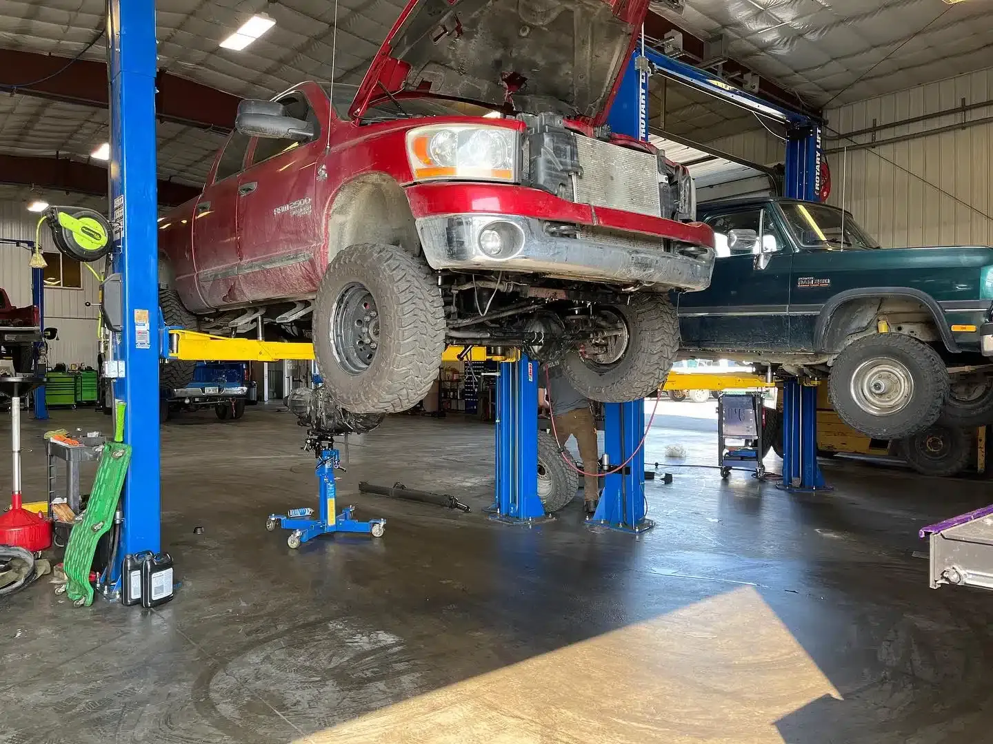 A red pickup truck is elevated on a blue hydraulic lift in a garage. The truck’s hood is open, and various tools and equipment are visible around the workshop. A second vehicle is lifted in the background.