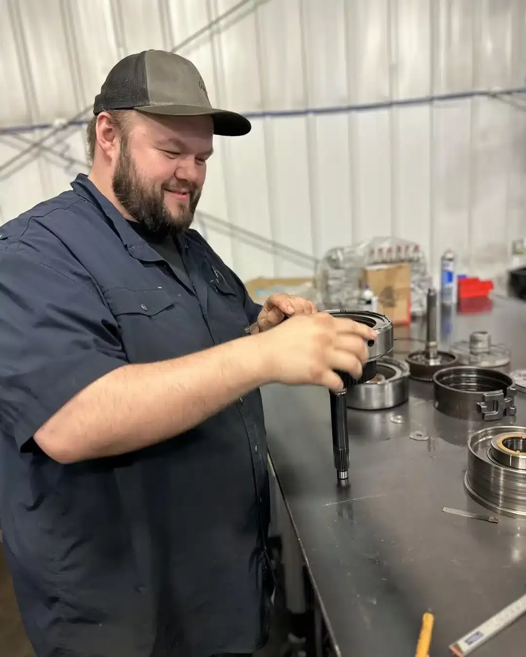 A bearded man in a dark cap and shirt works on an automotive part at a metal table in a workshop. He is holding a tool, surrounded by various mechanical components. The workshop has white walls and a metallic finish.